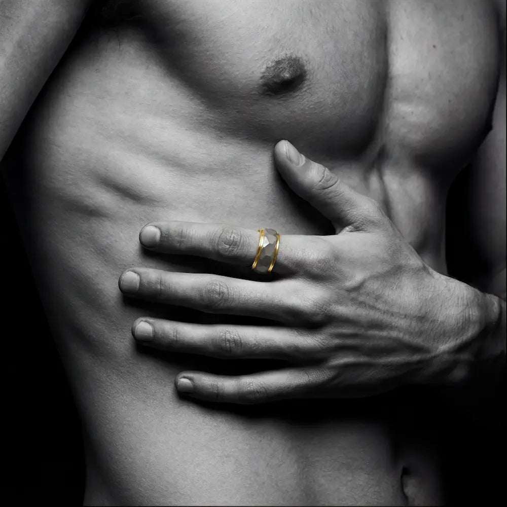 Man's hand wearing The Prismatic Pillar Aerospace-grade Tungsten ring resting firmly against a sculpted, muscular torso in a dramatic black-and-white aesthetic, with the gold accents highlighted as the sole color anchor.