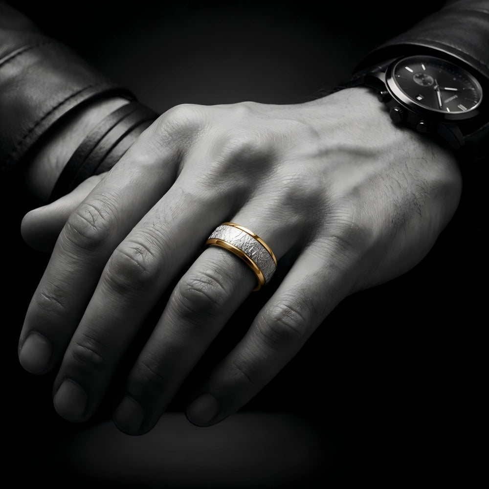 Man's hand wearing The Astral Pathway ring featuring 18K Gold PVD coating on Aerospace-grade Tungsten with an Authentic Muonionalusta Meteorite inlay, resting against a dark environment with a leather cuff and chronograph timepiece.