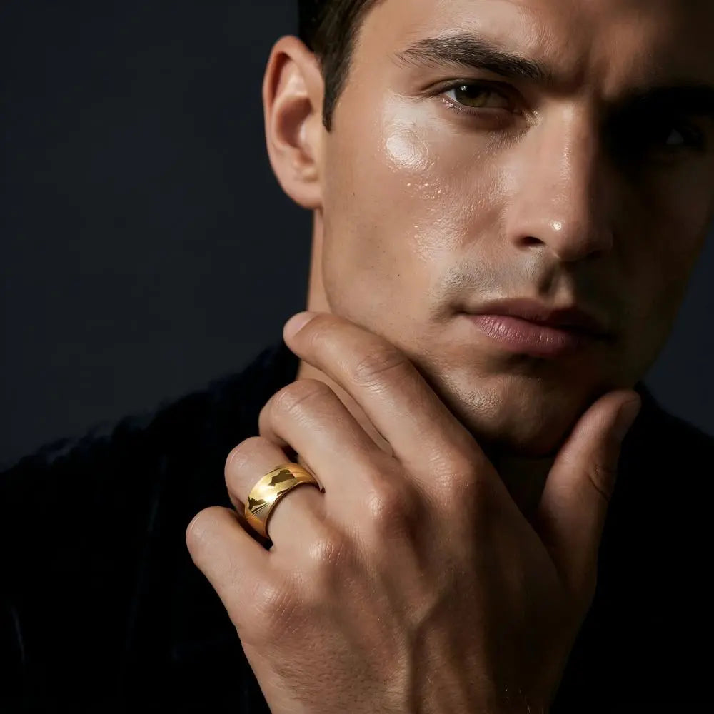 Man's hand wearing The Altar Strata 18K Gold PVD Forged Damascus steel ring, hand raised with fingers lightly touching chin in a contemplative pose against a dark background.