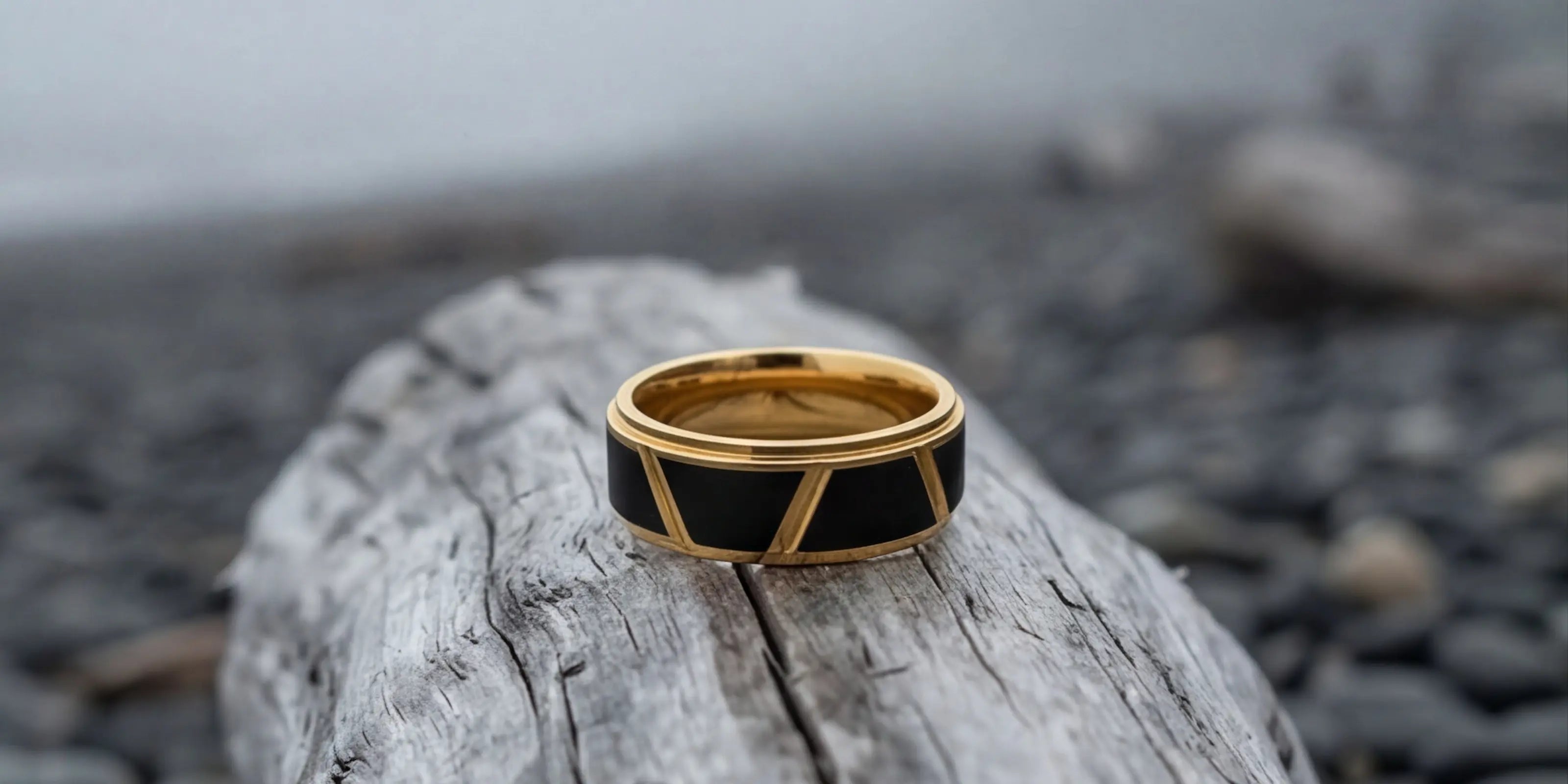 The Linear Divide Precision-engineered ring featuring black segmented geometric panels and an 18K Gold PVD coated tungsten base, resting on a weathered log along a misty, rocky shoreline.