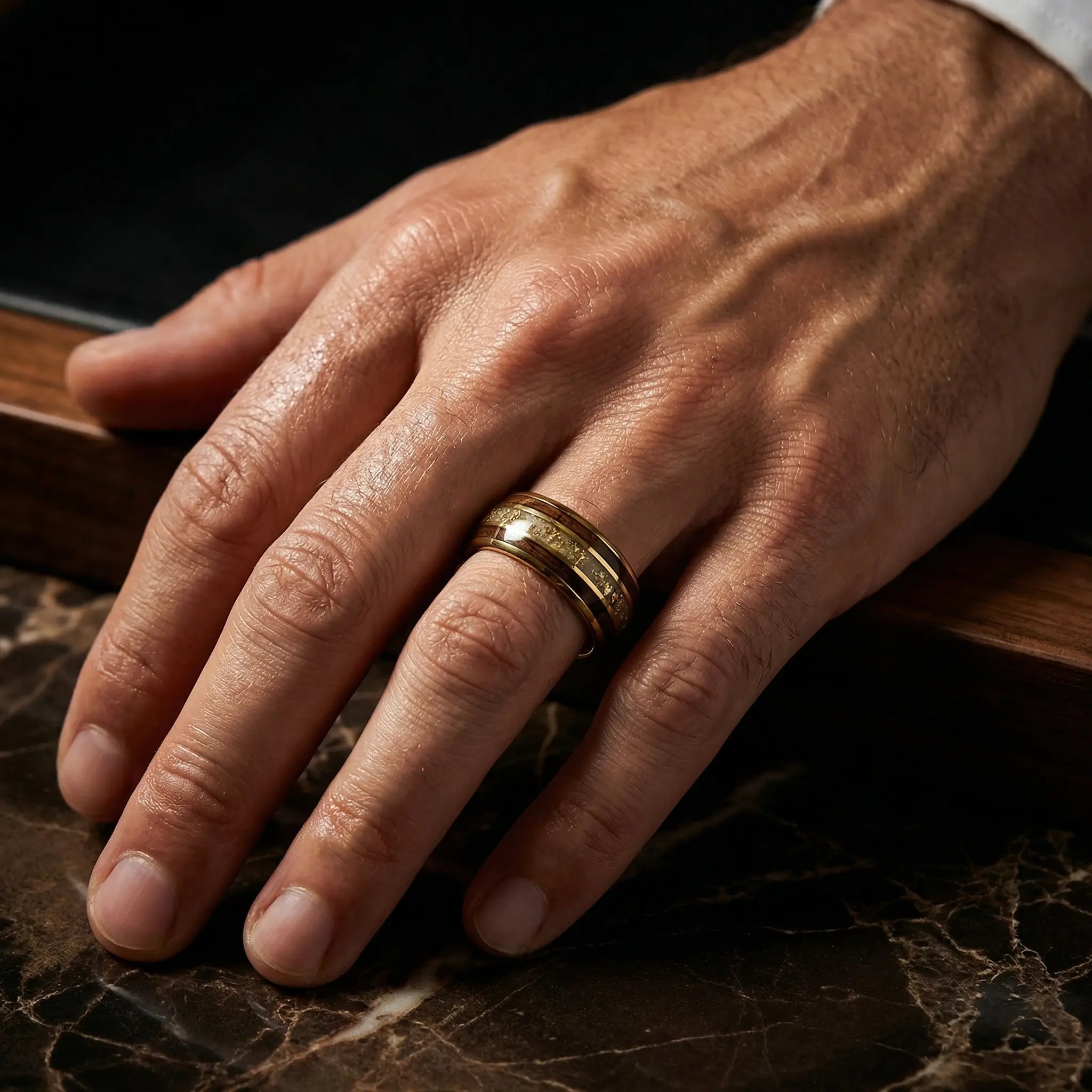 Man's hand wearing The Chronos Vessel 18K Gold PVD Tungsten ring resting on a polished dark marble tabletop bordered by rich hardwood.