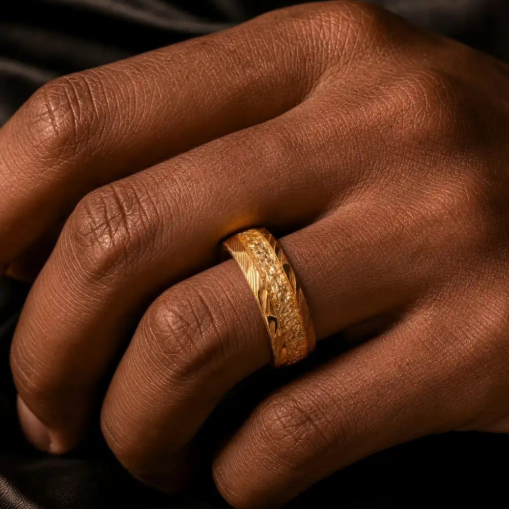 Man's hand wearing The Bedrock Crucible 18K Gold PVD Forged Damascus steel ring resting against a dark structured background.