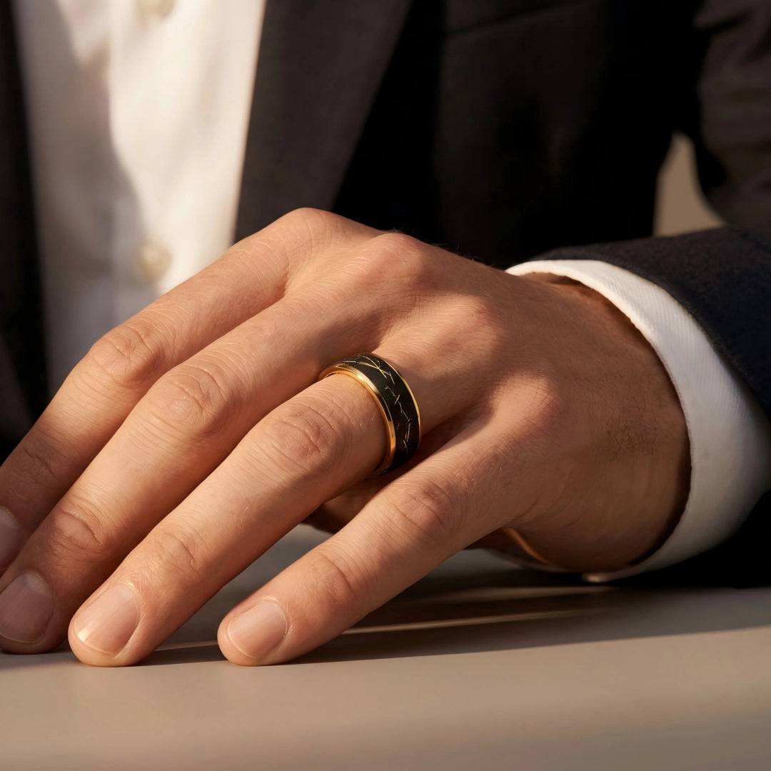 Man's hand wearing The Aureate Thread 18K Gold PVD ring resting on a sleek table surface with a dark tailored suit cuff.